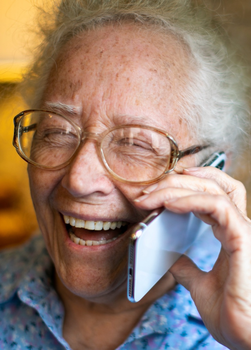 An older woman wearing glasses, laughing during a phone call.